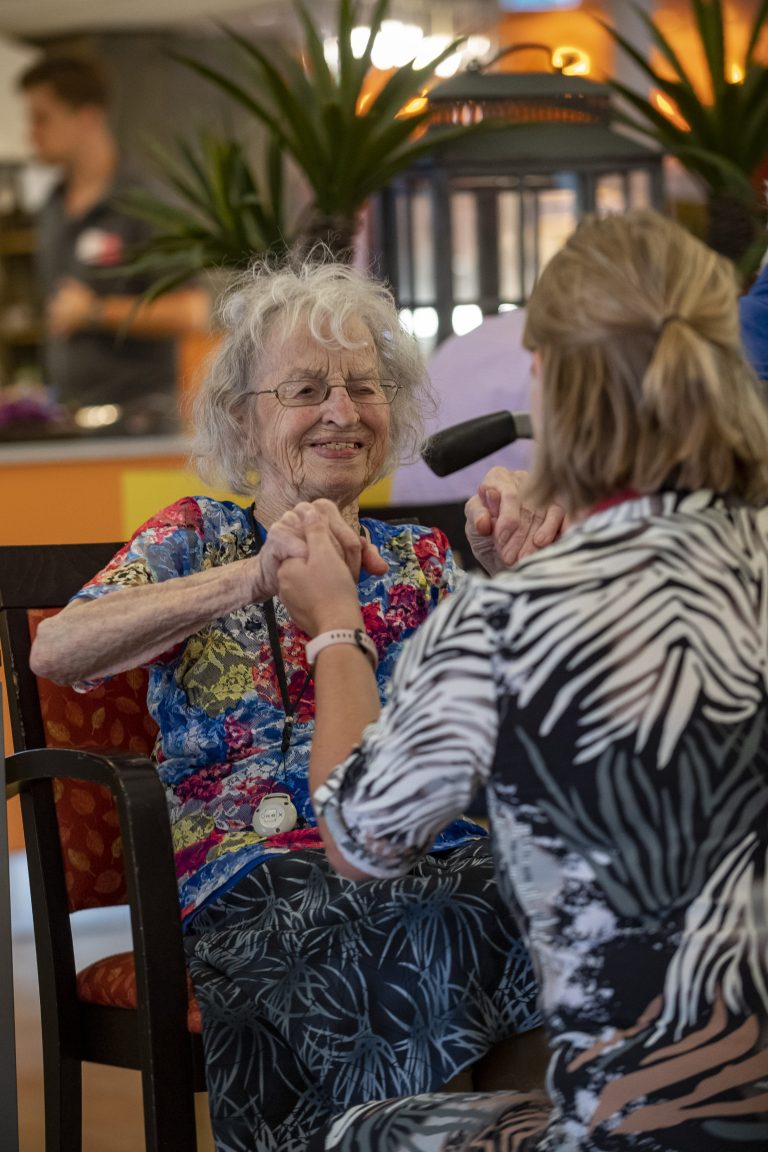 Een bewoonster van een verzorgingshuis van tanteLouise zit in haar rolstoel en houdt glimlachend de handen vast van een medewerker.